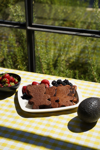 A children's plate of Spotty Choc Avocado Gluten Free and Date-Sweetened Pancakes in the shape of bears topped with fresh strawberries and blackberries on a yellow-and-white checkered tablecloth, with an avocado and a bowl of strawberries nearby, set in front of a window with a blurred garden background.