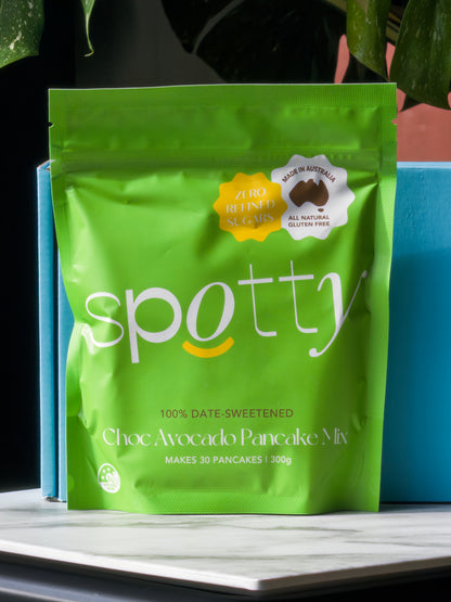 A bright green stand-up pouch of Spotty Choc Avocado Gluten Free and Date-Sweetened Pancake Mix, displayed on a white marble surface with a blue box behind it and dark green leaves at the top.