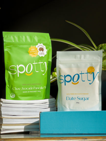 Two Spotty brand pouches of the Choc Avocado Pancake Mix and Date Sugar displayed on a shelf, with one green pouch on a stack of books and one white pouch next to a blue box. Gluten Free and Date-Sweetened.