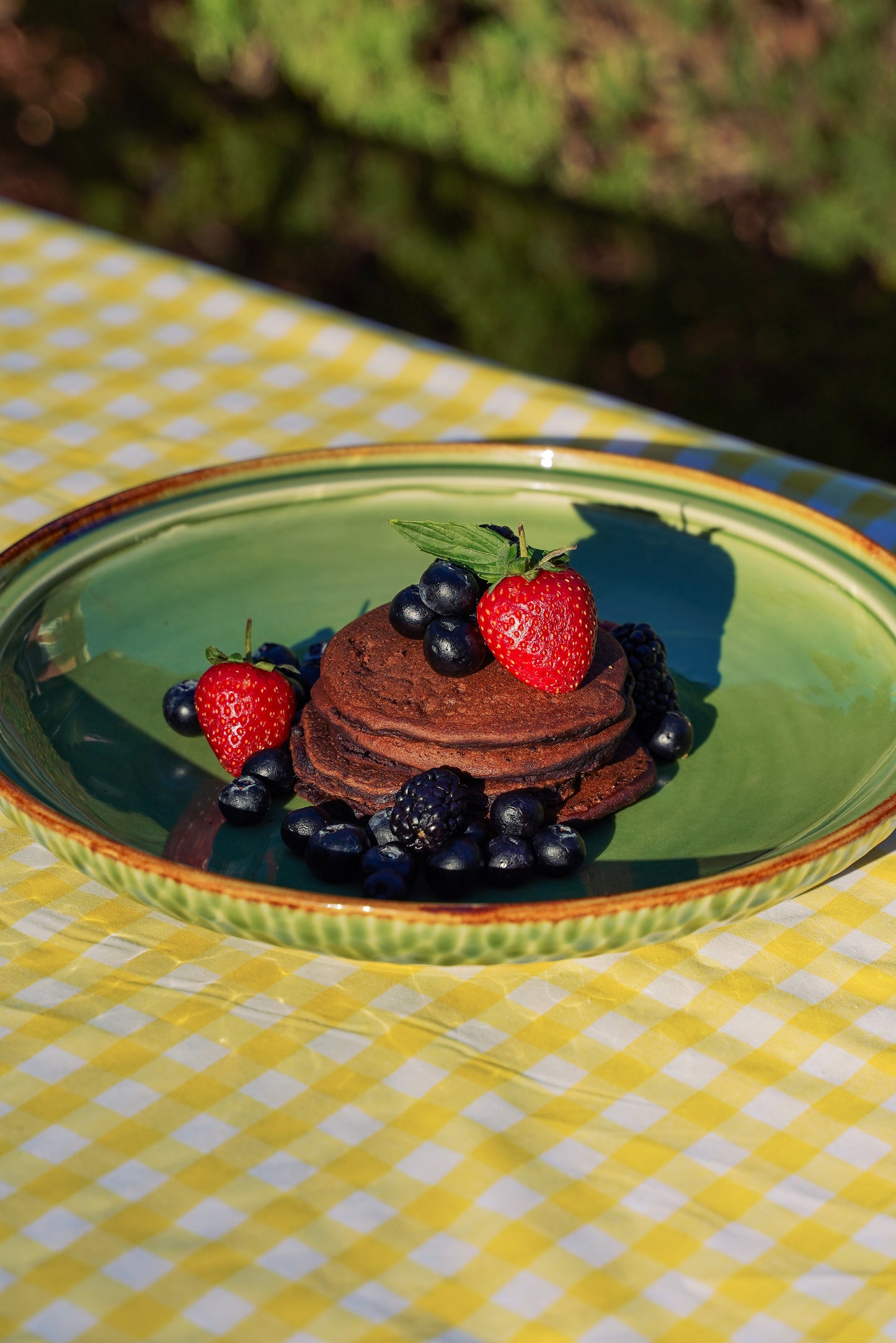 A stack of Spotty Choc Avocado Gluten Free and Date-Sweetened Pancakes topped with fresh strawberries, blueberries, and blackberries, served on a green ceramic plate over a yellow and white checkered tablecloth outdoors.
